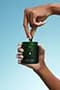 Hand dropping a probiotic capsule into a small green glass Seed container against a blue background.